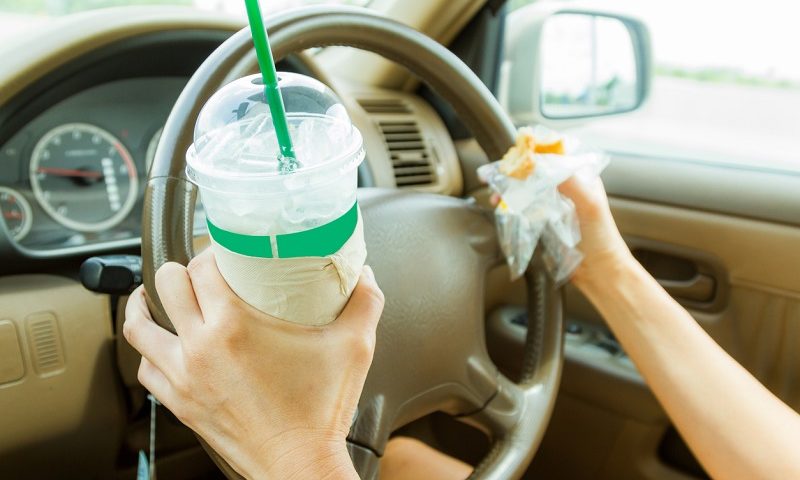 Woman driving a car on the road with a drink and burger in her hands.